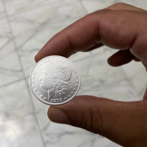 May include: A silver coin with a profile of a woman and the year 1878. The coin is held between two fingers, showcasing its intricate design and metallic sheen. The coin's edge features a detailed pattern.