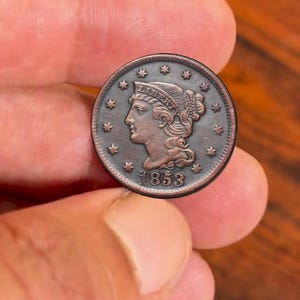 Może przedstawiać: Miedziana moneta z 1853 roku, przedstawiająca profil kobiety ze słowem "LIBERTY" nad głową. Moneta otoczona jest gwiazdami i ozdobnym obramowaniem. Moneta trzymana jest między dwoma palcami.