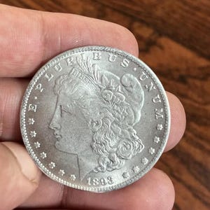 May include: A silver coin with the profile of a woman, the text "LIBERTY" and the year "1893". The coin has a detailed design and is held in a hand. The coin's edge has a star pattern.