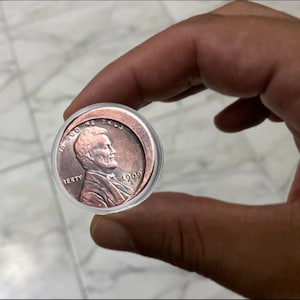 May include: A copper-colored 1909 penny coin in a clear plastic case. The coin features a profile of a man and the words "IN GOD WE TRUST" and "LIBERTY". The year "1909" is also visible.