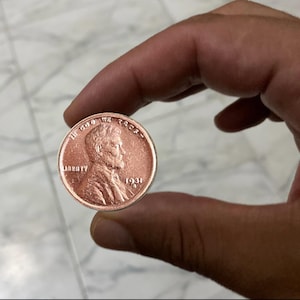 May include: A copper-colored 1931 penny held between fingers. The coin features a profile of a man and the word "LIBERTY". The text "IN GOD WE TRUST" and the year "1931" are also visible.