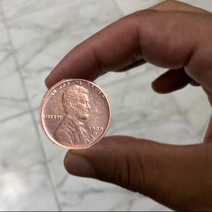 May include: A copper-colored penny coin from 1909, held between fingers. The coin features a profile of a man and the word "LIBERTY". The text "IN GOD WE TRUST" is also visible.