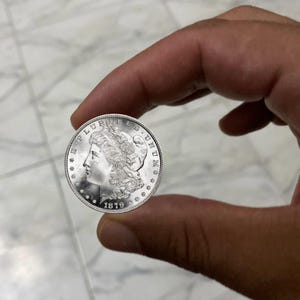 May include: A silver coin with a profile of a woman and the text "E PLURIBUS UNUM" and the date "1879". The coin is held between fingers.