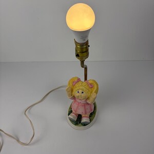 May include: A table lamp featuring a figurine of a blonde doll with pigtails and pink bows, wearing a pink polka-dot dress. The doll sits on a green base, supporting a brass-colored metal stem with a single light bulb socket and a glowing light bulb.