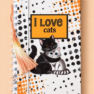 May include: A spiral-bound notebook with a white cover featuring black and orange polka dots, a rectangular orange box with the text I Love cats, and an illustration of a black cat hugging a grey tabby cat, finished with an orange tassel.