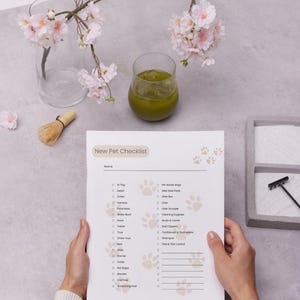 The image shows a "New Pet Checklist" held by a person, listing essential pet care items such as a tag, leash, collar, harness, food and water bowls, treats, toys, bedding, cleaning supplies, grooming tools, and health essentials. The checklist is on a clipboard with surrounding objects including a glass of green liquid, a branch with pink flowers, a whisk, and a tray with sand and a rake.