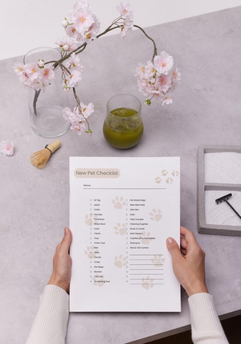 The image shows a "New Pet Checklist" held by a person, listing essential pet care items such as a tag, leash, collar, harness, food and water bowls, treats, toys, bedding, cleaning supplies, grooming tools, and health essentials. The checklist is on a clipboard with surrounding objects including a glass of green liquid, a branch with pink flowers, a whisk, and a tray with sand and a rake.