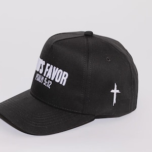 May include: Black baseball cap with the text "GOD'S FAVOR" and "PSALM 5:12" embroidered in white. A white cross is embroidered on the side. The cap has a curved brim and a button on top.