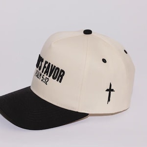 Op de afbeelding: Crèmekleurige baseballcap met een zwarte klep. De cap heeft de tekst "GOD'S FAVOR" en "PSALM 5:12" in zwarte letters. Een zwart kruis staat op de zijkant.