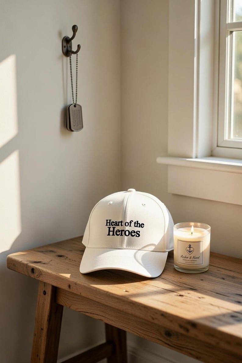 May include: A white baseball cap with the text "Heart of the Heroes" in black lettering. A lit candle in a glass jar and a pair of dog tags hang on the wall. The items are on a wooden bench.