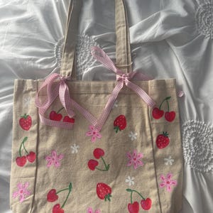 May include: A beige canvas tote bag featuring hand-painted red strawberries, red cherries, and pink flowers. The bag includes two shoulder straps and is decorated with a light pink and white gingham ribbon tied in bows at the base of the straps.