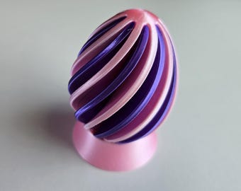 Op de afbeelding: Een decoratief eivormig object met een spiraalvormig ontwerp. Het ei heeft afwisselende banden van roze en paars en is gemonteerd op een roze cirkelvormige basis. Het object is waarschijnlijk 3D-geprint en heeft een gladde, glanzende afwerking.