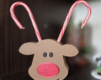 Op de afbeelding: Een bruine rendiervormige houder met een rode neus en zwarte ogen, met twee zuurstokken. De zuurstokken zijn rood en wit gestreept en buigen omhoog, wat op geweien lijkt. Een feestelijke kerstversiering.