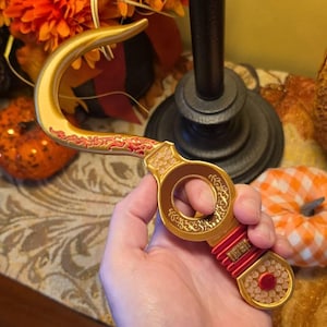 May include: A gold-colored hook prop featuring a curved blade with intricate red detailing, a circular handle with ornate engravings, a red wrapped grip, and a circular pommel with a central red accent, designed with a fantasy aesthetic.