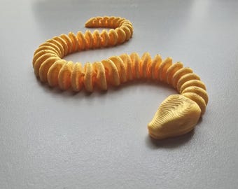 Op de afbeelding: Een gele, 3D-geprinte, beweeglijke slangenknuffel. De slang bestaat uit segmenten, waardoor hij flexibel kan bewegen, en heeft een gedetailleerde kop. Het speelgoed is ontworpen om op een echte slang te lijken.