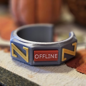 May include: A gray, 3D-printed bracelet with gold-colored 'Z' designs on each side. A red rectangle in the center displays the word "OFFLINE" in white. The bracelet is on a patterned surface.