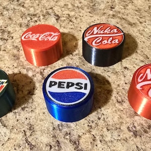 May include: Five cylindrical bottle stoppers featuring logos for Coca-Cola, Nuka Cola, Mountain Dew, and Pepsi. The stoppers are red, black, blue, and green. The logos are white and red.