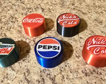 Op de afbeelding: Vijf cilindrische flesstoppers met logo's voor Coca-Cola, Nuka Cola, Mountain Dew en Pepsi. De stoppers zijn rood, zwart, blauw en groen. De logo's zijn wit en rood.