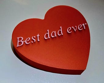 Op de afbeelding: Rode hartvormige doos met de tekst "Best dad ever" in witte letters. De doos heeft een licht verhoogde rand en een glad oppervlak. De tekst staat in het midden van het hart.