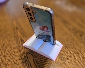 Op de afbeelding: Een smartphone staat in een roze en witte boekenstandaard. De telefoon heeft een doorzichtige hoes met een cartoonkarakterafbeelding. De standaard staat op een houten oppervlak.