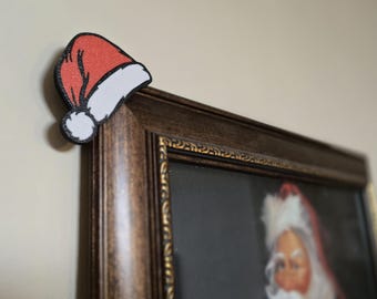 Op de afbeelding: Een feestelijke kerstversiering met een rood-witte kerstmuts, geplaatst op de hoek van een ingelijst portret. De hoed heeft een zwarte omtrek en een witte pompon. De lijst is donkerbruin.