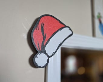 Op de afbeelding: Een rode en witte kerstmuts met een zwarte omtrek. De hoed is gemaakt van een textuurrijk materiaal en heeft een witte pompon. Een kerstversiering.