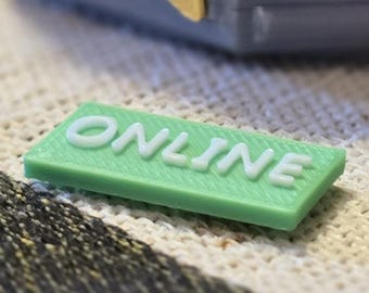 Op de afbeelding: Een klein, rechthoekig, lichtgroen object met het woord "ONLINE" in witte, verhoogde letters. Het object ligt op een gestructureerd, gebroken wit en grijs oppervlak. De achtergrond bevat een deel van een grijs en goudkleurig object.
