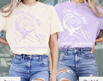 May include: Two short-sleeve crew neck t-shirts in ivory and orchid colors. Each shirt features a graphic of a person with a towel on their head and cucumber slices on their eyes, with the text Girls Gone Mild, Lauren's Bachelorette, New York 2025.