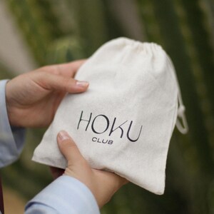 以下が含まれることがあります： 黒色の「HOKU CLUB」の文字が入った、クリーム色の巾着袋。ぼやけた緑色の背景の中で、人が袋を持っています。