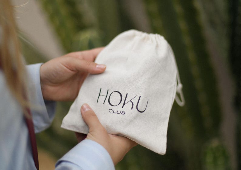 以下が含まれることがあります： 黒色の「HOKU CLUB」の文字が入った、クリーム色の巾着袋。ぼやけた緑色の背景の中で、人が袋を持っています。