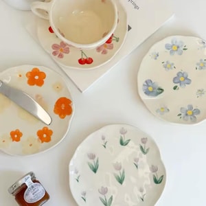 May include: Four ceramic plates with floral designs. The plates feature different floral patterns, including orange and yellow flowers, blue flowers, and pink and white tulips. The plates are all white with a slightly irregular shape.