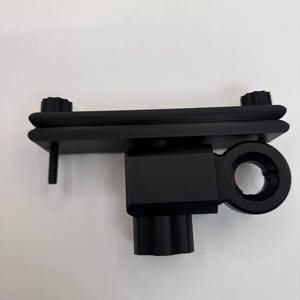 May include: A black metal device with a rectangular base, a cylindrical post, and a circular opening. Two black knobs are on top, and a screw extends from the side. The device is designed for mounting or securing.