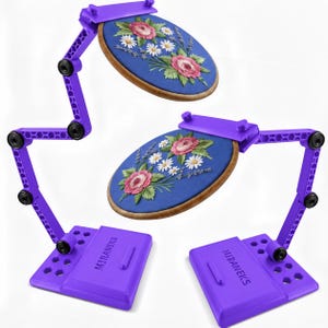 May include: Two purple adjustable embroidery hoop stands with articulated arms and rectangular bases labeled MIRANEKS. Each stand holds a wooden embroidery hoop containing blue fabric with a floral design of pink roses and white daisies.