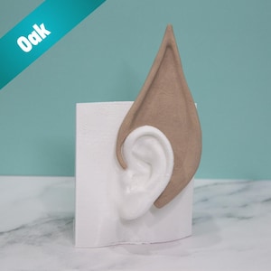 May include: A pair of prosthetic elf ears in a light brown color, with a white ear insert, displayed on a white rectangular base. The word "Oak" is printed on a teal diagonal banner.