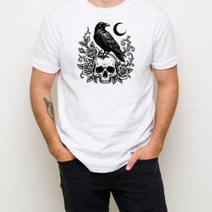 Puede incluir: Una camiseta blanca de manga corta con un estampado gr&aacute;fico negro en el pecho. El dise&ntilde;o muestra un cuervo posado sobre un cr&aacute;neo humano, rodeado de rosas en flor, enredaderas espinosas y una luna creciente en una ilustraci&oacute;n detallada.