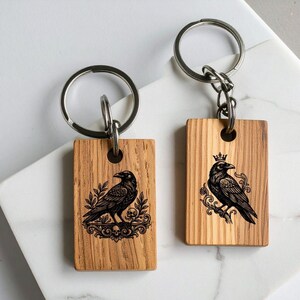 Puede incluir: Dos llaveros rectangulares de madera con dise&ntilde;os grabados a l&aacute;ser en negro. El llavero izquierdo muestra un cuervo sobre follaje ornamentado con calaveras, el derecho un cuervo coronado sobre una rama con volutas decorativas.