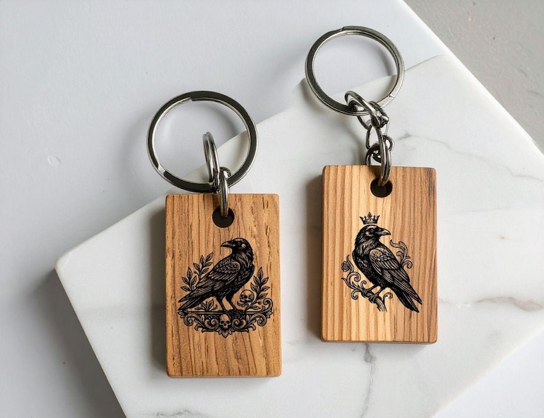 Puede incluir: Dos llaveros rectangulares de madera con dise&ntilde;os grabados a l&aacute;ser en negro. El llavero izquierdo muestra un cuervo sobre follaje ornamentado con calaveras, el derecho un cuervo coronado sobre una rama con volutas decorativas.