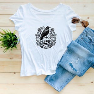 Puede incluir: Una camiseta blanca con cuello en V que presenta un estampado gr&aacute;fico en blanco y negro de un cuervo posado sobre una calavera, rodeado de enredaderas de rosas espinosas. La camiseta se combina con vaqueros azules y gafas de sol.