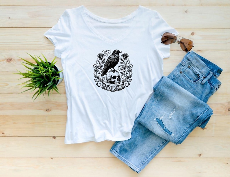 Puede incluir: Una camiseta blanca con cuello en V que presenta un estampado gr&aacute;fico en blanco y negro de un cuervo posado sobre una calavera, rodeado de enredaderas de rosas espinosas. La camiseta se combina con vaqueros azules y gafas de sol.
