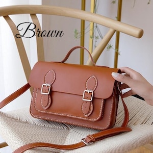 Może przedstawiać: Brązowa torba listonoszka z górnym uchwytem i długim, regulowanym paskiem. Torba posiada dwie srebrne klamry i białe detale szwów. Słowo "Brown" jest napisane kursywą w lewym górnym rogu.