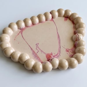 May include: A decorative, heart-shaped ceramic tray with a light beige base and a border of round, beige beads. The tray's surface features a marbled design in shades of pink and red. The tray is ideal for displaying small items.
