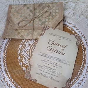May include: A woven envelope with a twine tie sits on a burlap placemat with a white lace design. A wedding invitation with ornate borders and text is also on the placemat. The invitation reads "Samuel at Katrina".