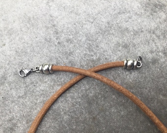 May include: Close-up of a tan leather cord necklace with silver-colored metal clasps. The cord is curved and crossed, with a lobster clasp on one end and a loop on the other.