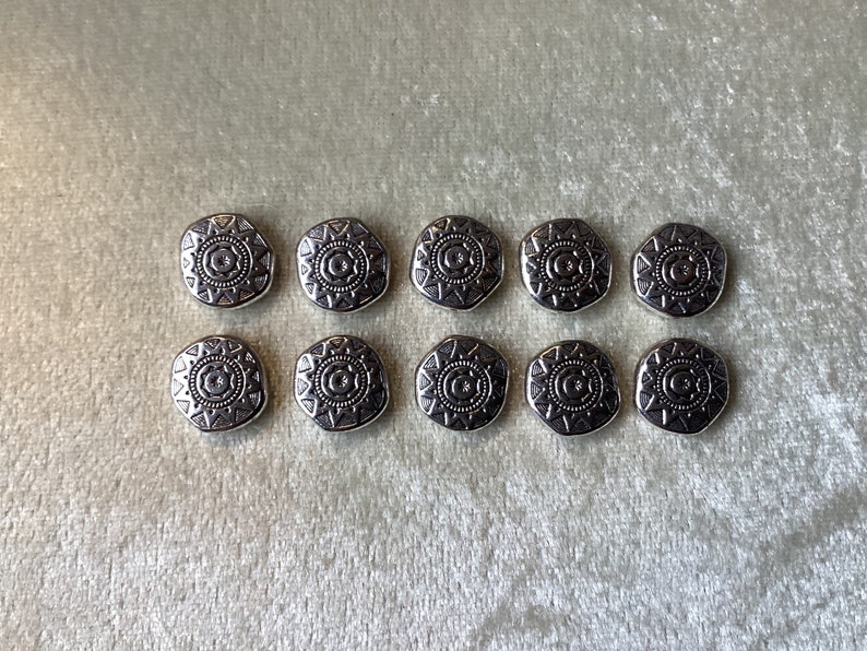 May include: Ten silver-toned buttons with a geometric design. Each button features a circular pattern with a starburst design.