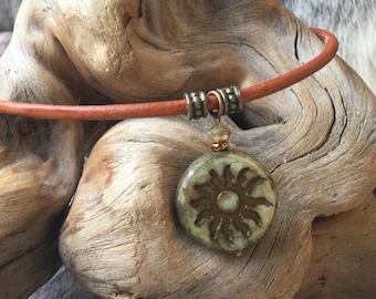 May include: A necklace featuring a round, mottled green pendant with a sun motif embossed in the center. The pendant hangs from a smooth, orange leather cord, accented by two metallic, cylindrical beads with a textured, dotted pattern.