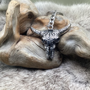 May include: A silver-colored bull skull keychain with a chain and key ring. The skull has detailed horns and a textured surface. The keychain is displayed on a piece of driftwood and a furry, gray surface.