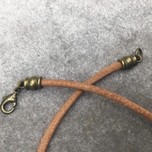 K&ouml;nnte beinhalten: Nahaufnahme einer braunen Lederschnur mit antik-messingfarbenen Metallverschl&uuml;ssen und einem Karabinerhaken. Die Schnur ist in der Mitte gekreuzt und zeigt die Details des Materials und der Hardware.
