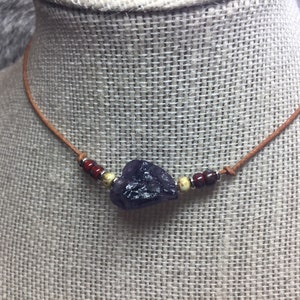 May include: A brown leather necklace with a dark purple gemstone pendant and small red and yellow beads.