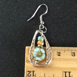 May include: Silver dangle earrings with a teardrop-shaped design. The earrings feature a blue and white flower bead, orange and green beads, and a silver hook.