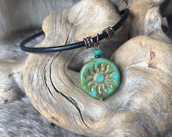 May include: A black leather choker necklace with a copper clasp and a turquoise pendant featuring a gold sun design.